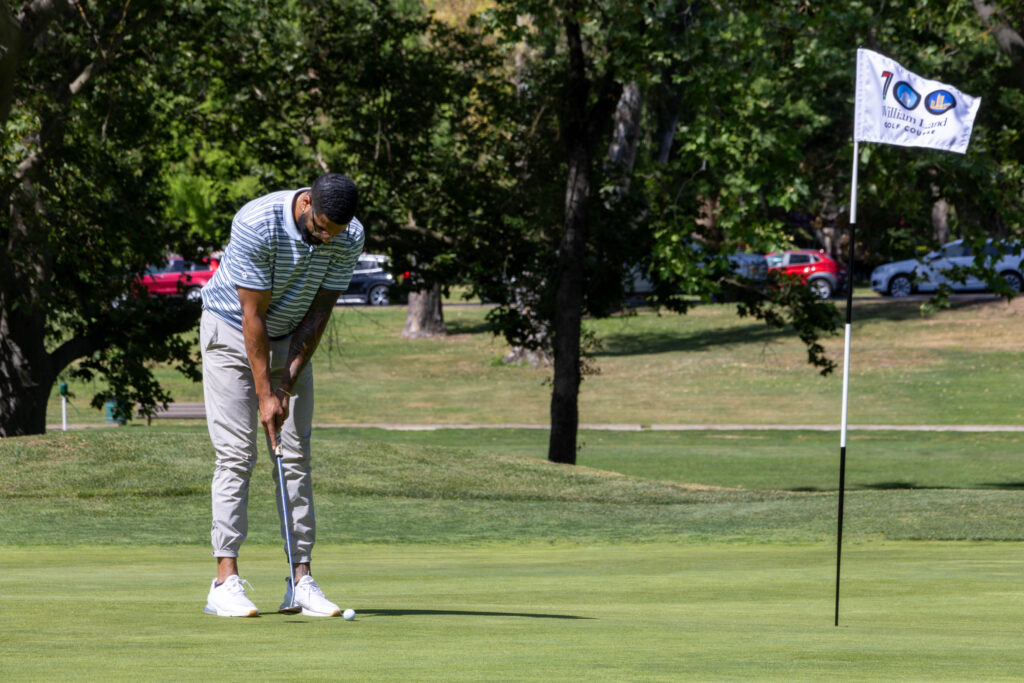 A single golfer is readying his putt on a green. He is standing several feet away from the pin with a special 100th anniversary William Land Golf Course logo on the flag.
