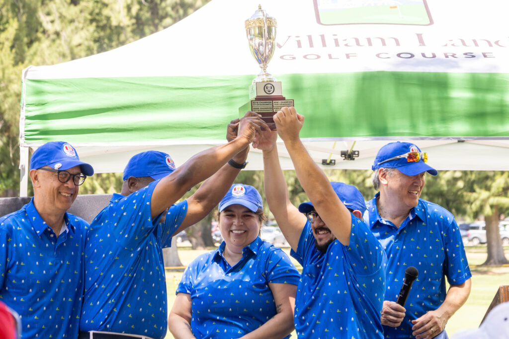 A group of city officials are holding up the Capitol Golf Challenge trophy and cheering. They are all wearing matching blue polos, commemorating the 100th anniversary of William Land.