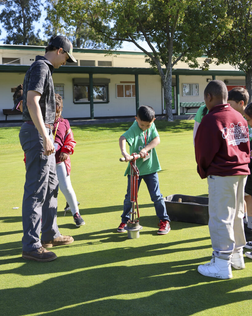 One student in a green shirt is attempting to cut a hole in a putting green with a tool while an adult supervises. Other students are standing around them and watching.