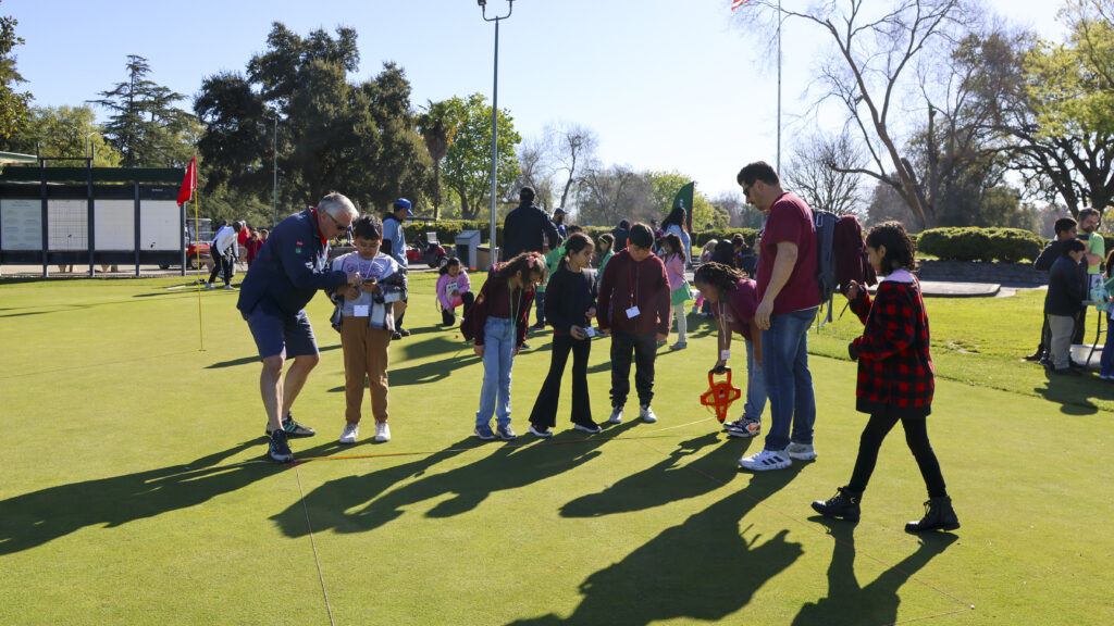 A group of students are standing in a line. One student stands at the end holding a large tape measure. An older man is holding the end of the tape measure at the other end of the students and showing one of them how to read it.