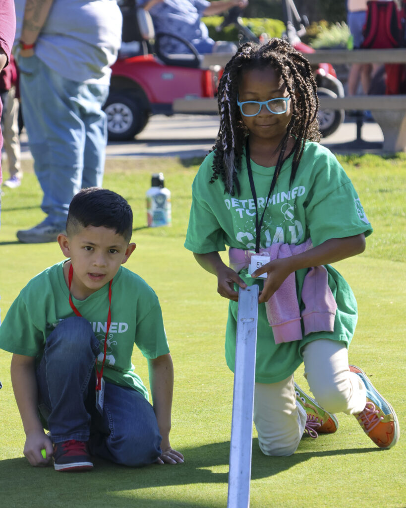 Two students are kneeling on the green. One is standing up slightly, holding a ramp used to calculate green speed after rolling a ball down it. The other is kneeling on the ground, watching the ball that rolled off the ramp.