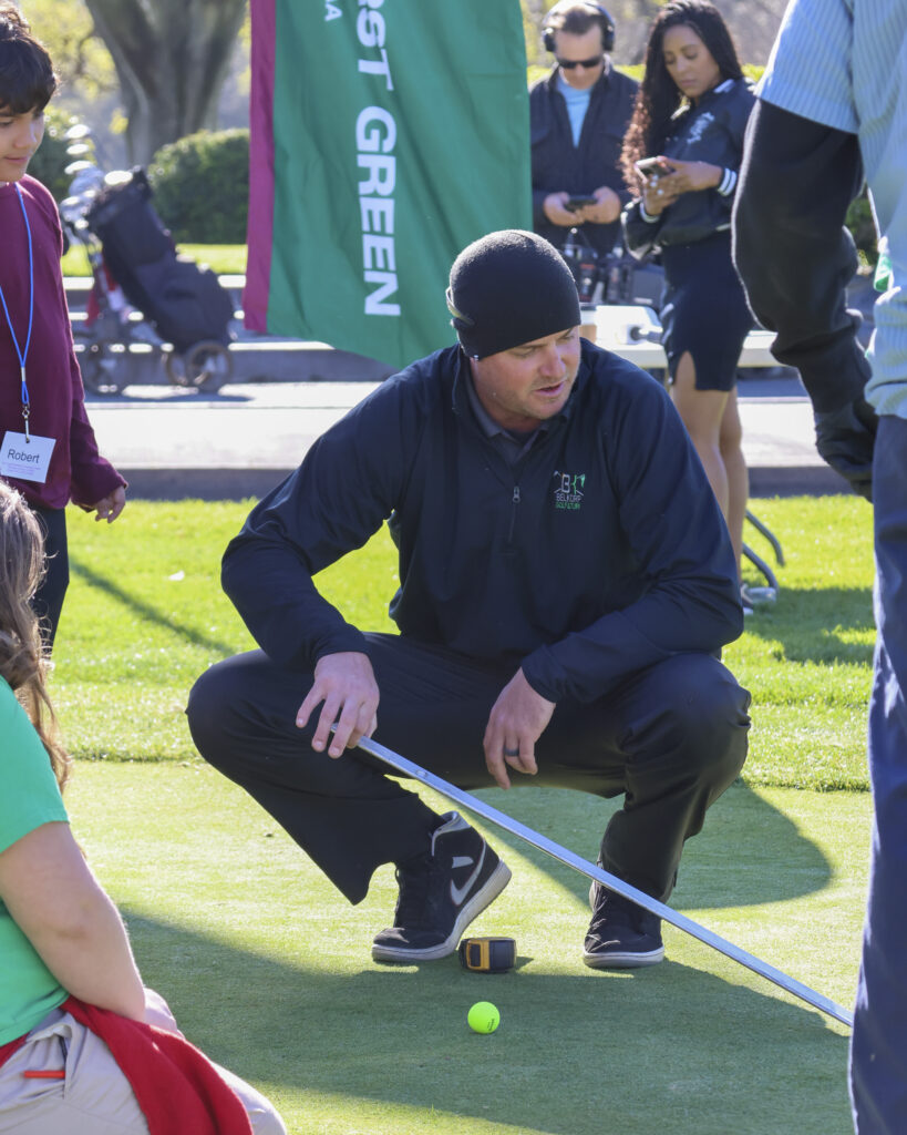 A man is squatting on a putting green, holding a ramp used to calculate green speed. He is demonstrating to a group of students how the ramp is used.