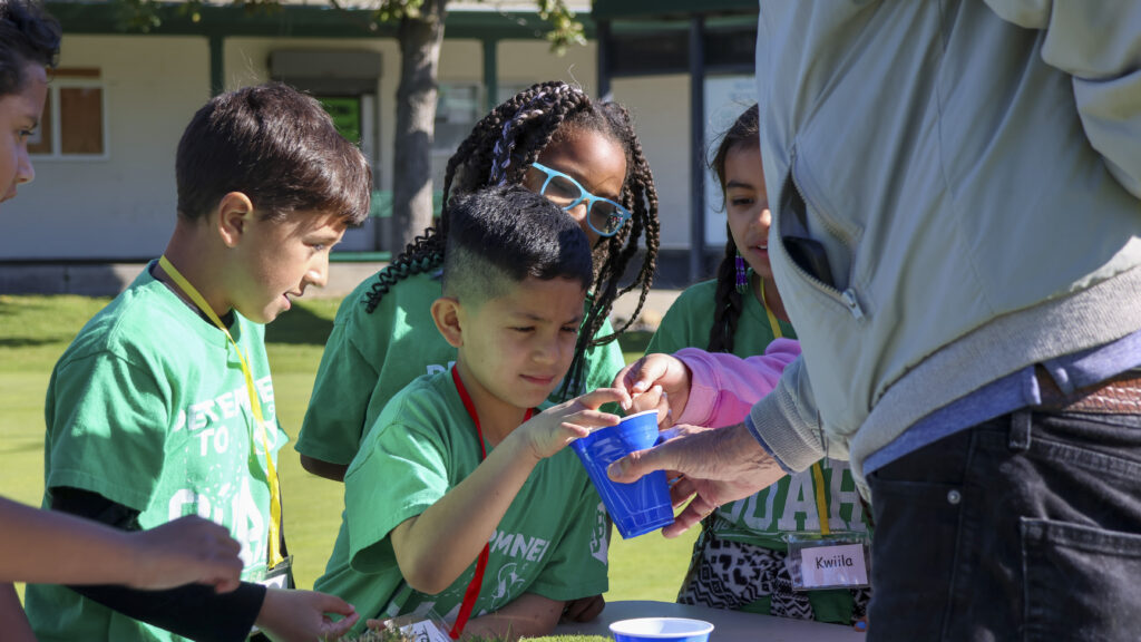 A group of students are gathering a man holding a blue cup. They are reaching into the cup, to inspect the seeds of grass inside.