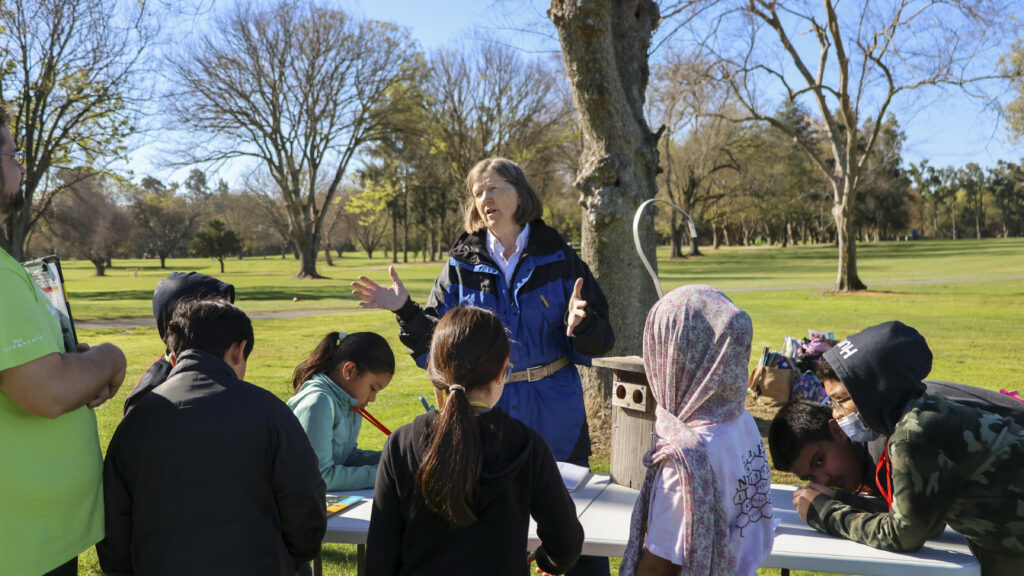 An older women in a blue jacket is talking to a group of students about wildlife preservation. On a table in front of her is a birdhouse with a large hook on top. 