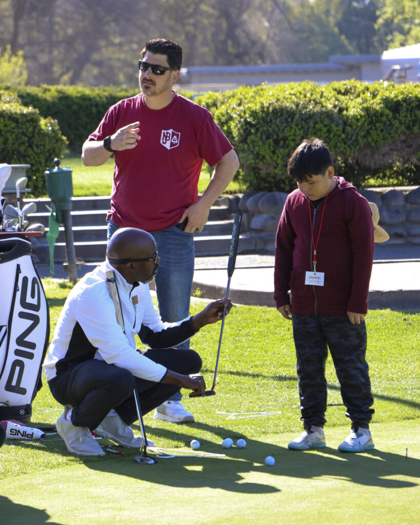 A man is kneeling next to a student on a putting green. He is holding up a putter for the student to use to hit several golf balls around them.