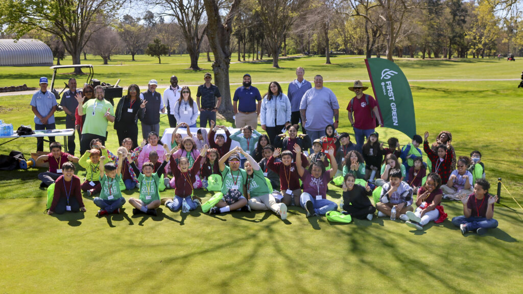 The students and the volunteers from the field trip are taking a group photo. The kids are sitting on the ground in front of the adults, in 3 rows, the adults are standing behind the kids. The kids are making silly poses and looking excited, while the adults are smiling.