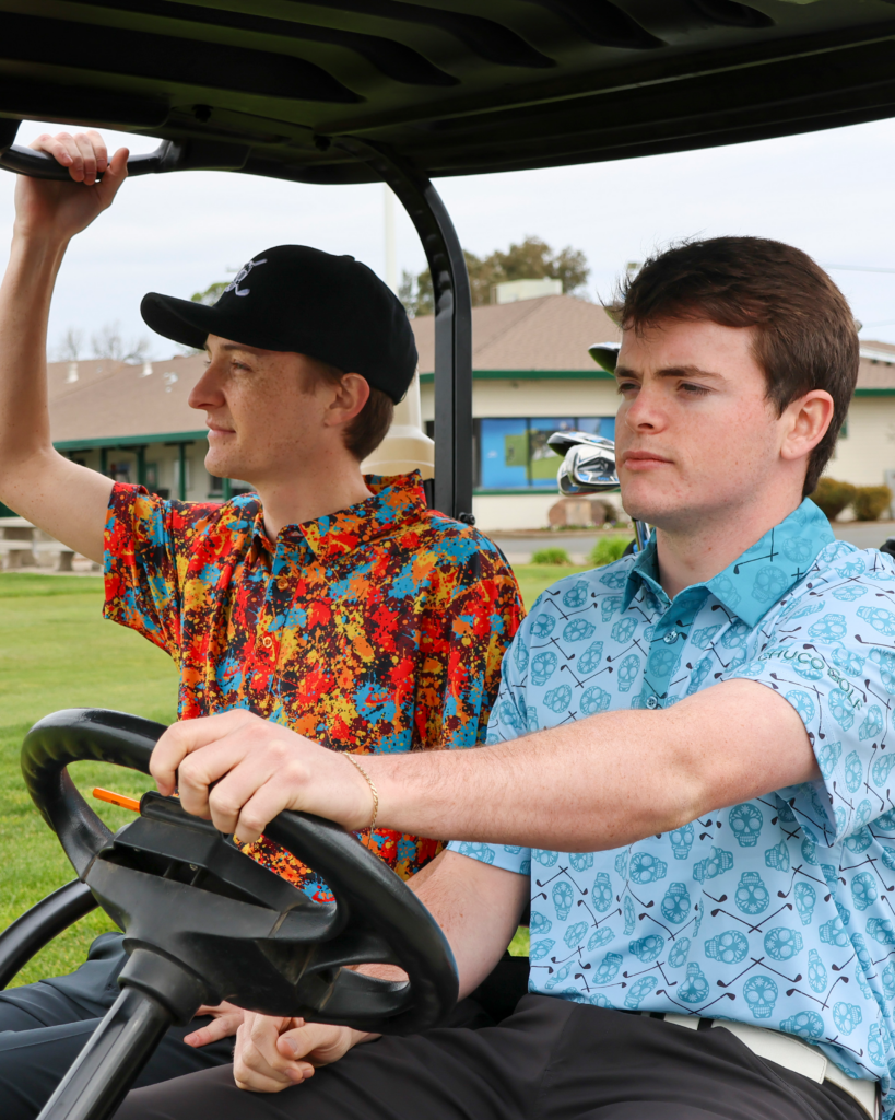 Two young men are sitting in a golf cart. The driver is holding the steering wheel while wearing a light blue polo with a sugar skull pattern. The passenger is holding the passenger assist grip. He is wearing a colorful polo shirt that looks like it's covered in splotches of red, orange, yellow, and light blue paint. He is wearing a black hat with the Chuco Golf logo, a white sugar skull with golf clubs.