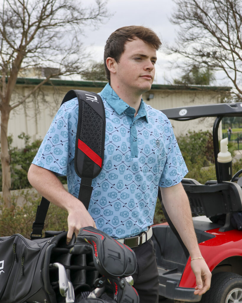 A young man is holding a golf bag to his side and walking away from a golf cart. He is wearing a light blue polo with a sugar skull pattern across it.
