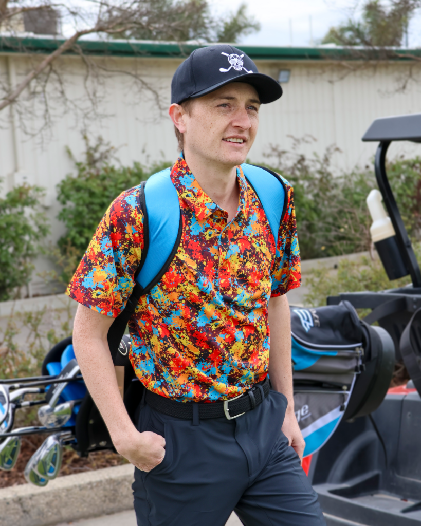 A young man is holding a golf bag on his back and walking away from a golf cart. He is wearing a colorful polo with. He is also wearing a black hat with the Chuco Golf logo, a white sugar skull with golf clubs.