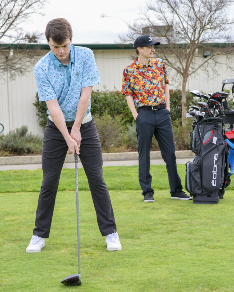 Two young men are standing on a tee box. One is in the foreground, addressing a golf ball on a tee. His face is focused on the ball. He is wearing a light blue polo with a sugar skull pattern across it. 'CHUCO GOLF' is emblazoned on his left sleeve. The second man is further back, looking right down the fairway. His hands are in his pockets and he wears a black hat with Chuco Golf's sugar skull logo. He is wearing a colorful polo shirt with a pattern that looks like red, blue, yellow, and orange paint splotches. To the right of him is a golf bag holding golf clubs.