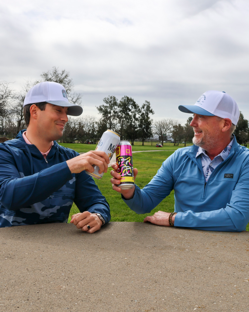 Two men are sitting at at stone table, making a toast with their beers. One holding a can of Modelo, wearing a blue hoodie partially covered with a camo pattern, a pink polo shirt, and a white TravisMathew hat. The other is holding a can of Gummy Worms and wearing a light blue jacket with no design on it, a blue and white polo with a leafy design, and wearing a different white TravisMathew hat with a blue lid.