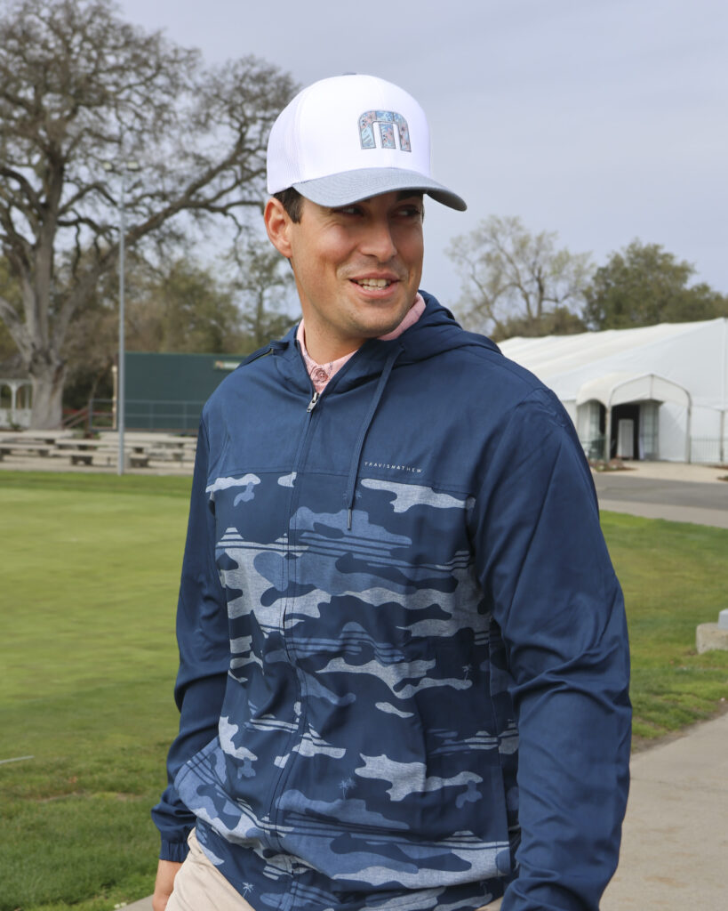 A man is walking in front of the practice green at Haggin Oaks. He is smiling and looking to his left, wearing a blue hoodie partially covered with a camo pattern, a pink polo shirt, and a white TravisMathew hat.