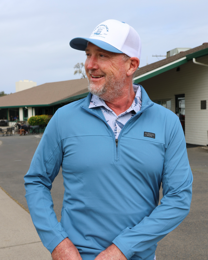 A mand standing outside the Haggin Oaks Golf Pro Shop. He is smiling and looking to his right while putting his sunglasses away in his pocket, wearing a light blue jacket with no design on it, a blue and white polo with a leafy design, and wearing a white TravisMathew hat with a blue lid.