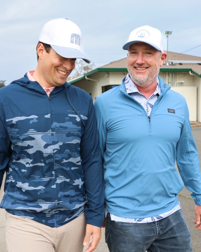 Two men standing outside the Haggin Oaks Golf Pro Shop, both wearing new TravisMathew. One is smiling and looking downward, wearing a blue hoodie partially covered with a camo pattern, a pink polo shirt, and a white TravisMathew hat. The other is smiling and looking at the camera wearing a light blue jacket with no design on it, a blue and white polo with a leafy design, and wearing a different white TravisMathew hat.