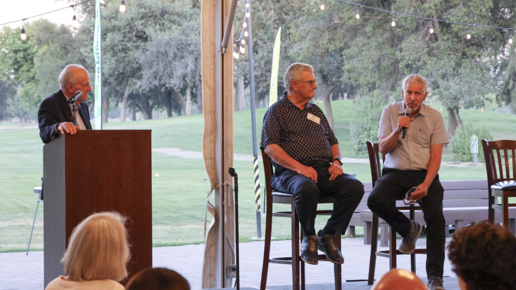 John Schumacher speaks into a microphone on stage after being inducted into the Hall of Fame. He is seated next to Ken Morton Sr. and Frank LaRosa.