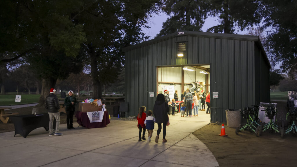 A wide shot of the William Land Golf Course cart barn at the beginning of the evening. Some families are inside talking with Santa. A table is set up outside for donations to the Sheriff Toy Project.
