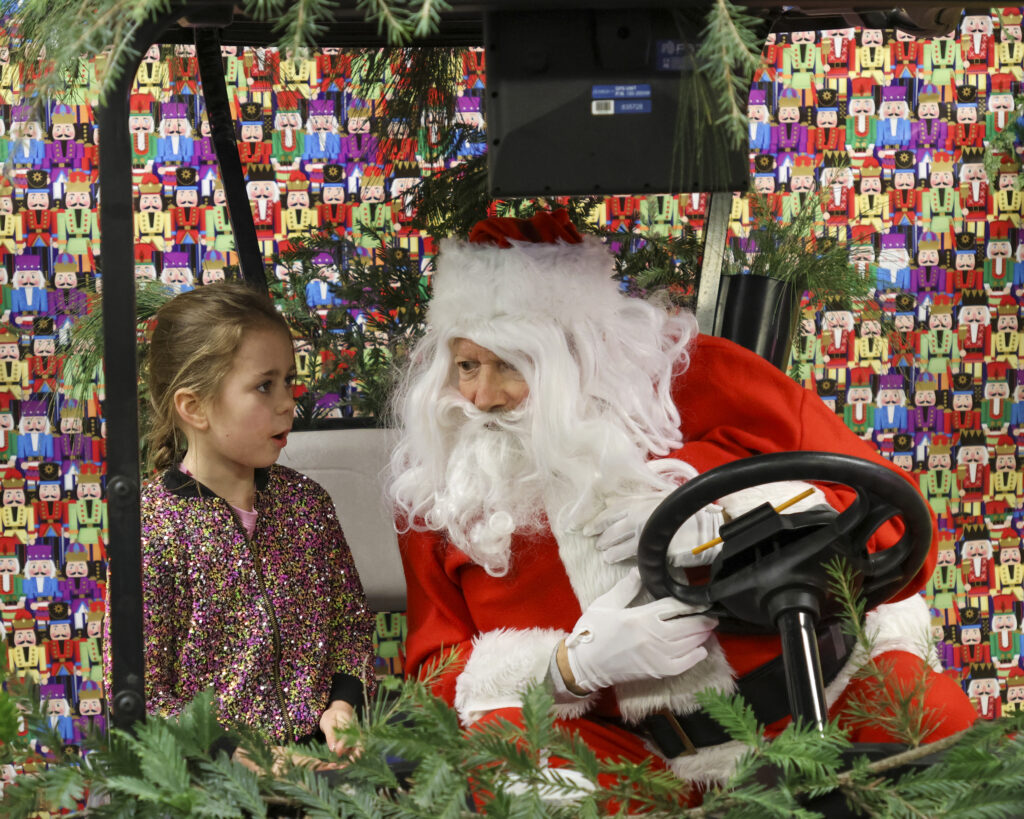Santa leans in toward a young girl sitting next to him as she tells him what she wants for Christmas.