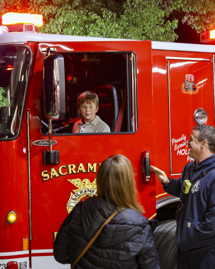 A young boy sits in the driver's seat of a firetruck while his mother and a fireman watch on.