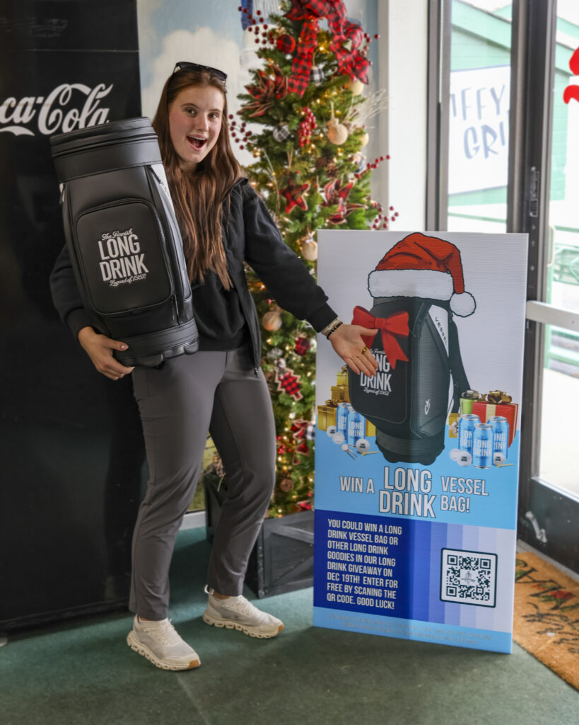 Soleil poses with the Long Drink Vessel bag next to the giveaway sign in the Pro Shop.