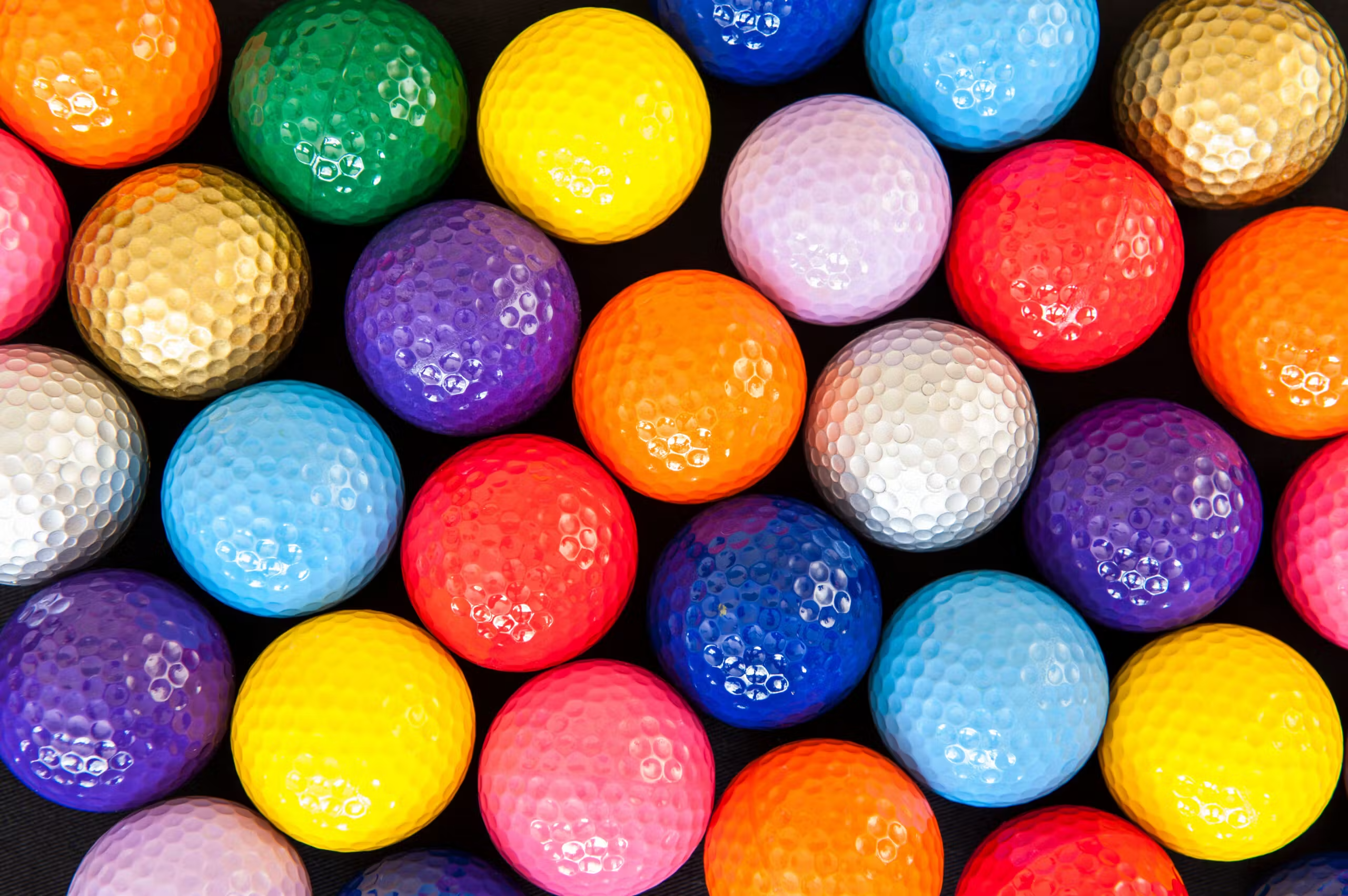An overhead photo of many golf balls of different colors lined up next to each other. They are red, blue, gold, orange, pink, purple, blue, white, yellow, silver, and more.