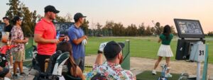 Golfers practicing at the Haggin Oaks driving range, with a woman hitting a shot while others watch from nearby bays with clubs and drinks at sunset