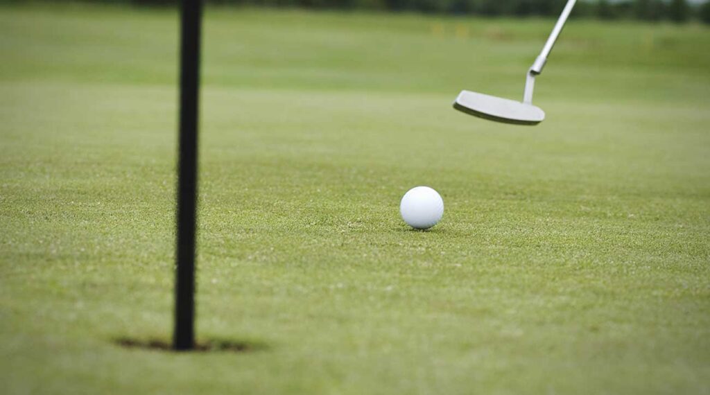 A close up of a ball rolling towards a golf hole with the flag stick in it. In the distance a putter can be seen, held off the ground after swing.