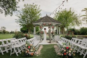 Outdoor wedding ceremony at Haggin Oaks with bride and groom standing beneath a floral-decorated gazebo, surrounded by white chairs and aisle arrangements on a manicured lawn.