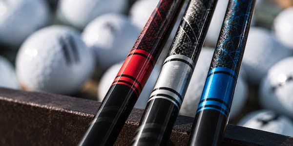 Close-up of three graphite golf shafts with red, silver, and blue accents and blurred golf balls in the background
